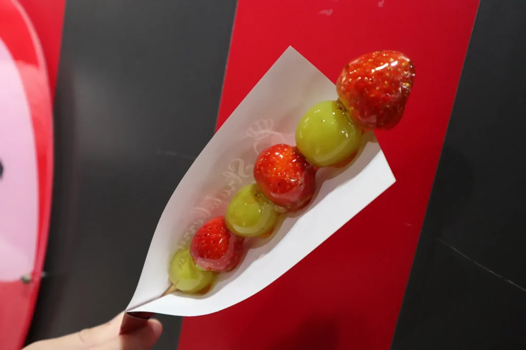 A skewer of candied strawberries and green grapes held in a paper tray